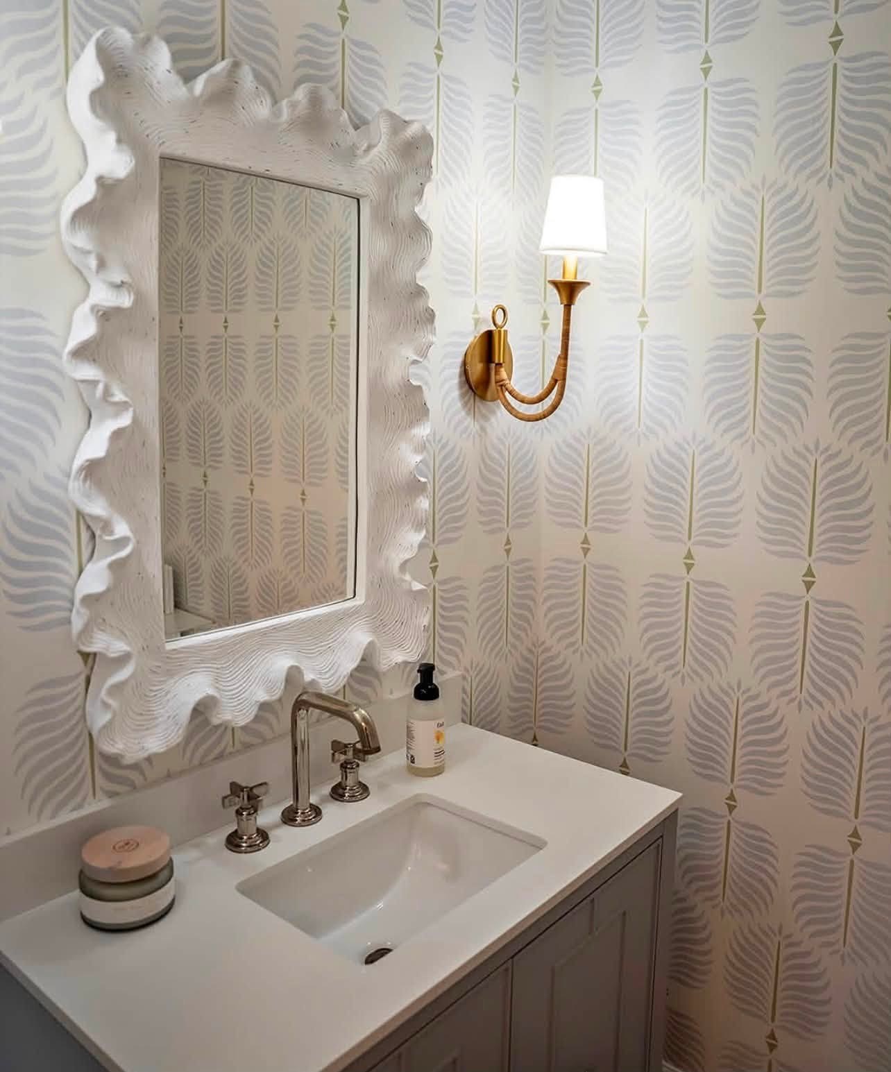 Soft blue leaf pattern wallpaper in a powder room with a white sculpted mirror and brass sconce installed by Jeff Handyman