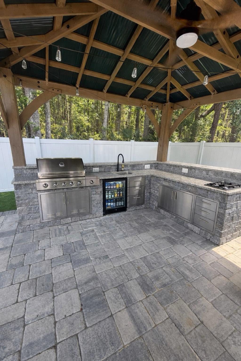 L-shaped outdoor kitchen under a wood pergola with built-in stainless grill, side cooktop, beverage cooler, stone counter, and paver patio