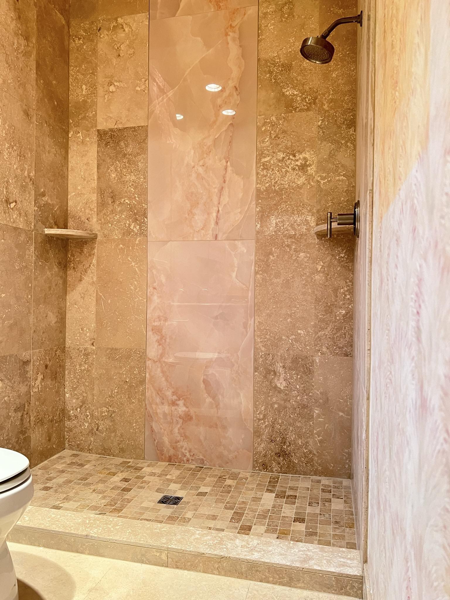 Walk-in shower with rose travertine tile walls, mosaic tile floor, and bronze rain shower head by Jeff Handyman