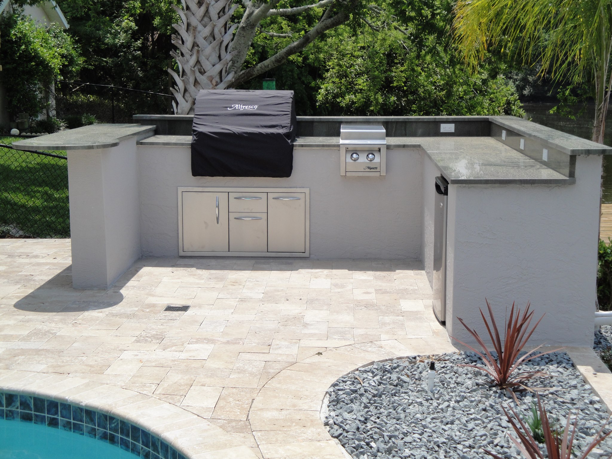 Pool-side U-shaped outdoor kitchen with travertine pavers, built-in Alfresco grill, stainless cabinets, and beverage fridge cut-out