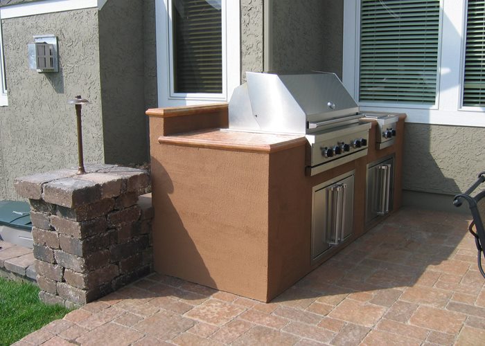 Stucco-front outdoor kitchen with copper-stained countertop, built-in stainless grill, and stone pillar accent on a paver patio